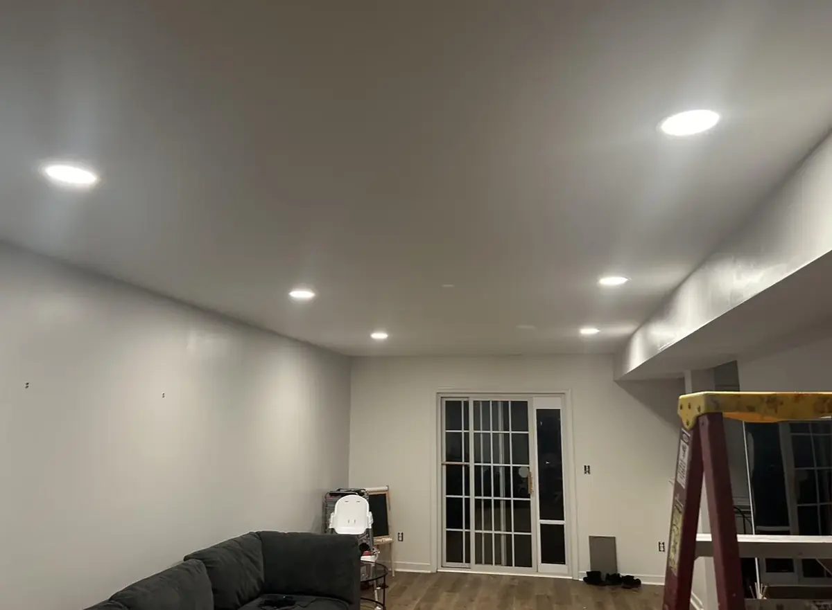 Grey living room ceiling with a row of recessed lights switched on, a sliding patio door, a sectional sofa and a stepladder still in the room