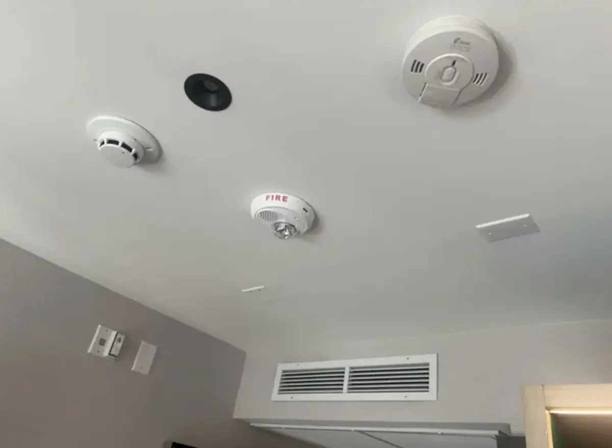 White ceiling with a smoke alarm, a second round detector, a red fire alarm horn-strobe and a black recessed light trim, with a wall vent below