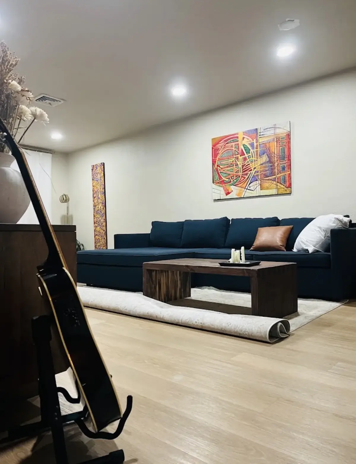 Family room with recessed ceiling lights, a navy sectional sofa, a wood coffee table, abstract paintings and a guitar on a stand in the foreground