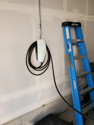Wall-mounted EV charger with its cable coiled on a hook, on unpainted garage drywall with conduit running up from it and a blue stepladder beside it