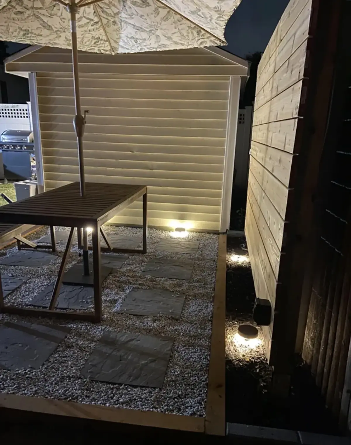 Backyard patio at night with low pathway lights along a gravel border, a wooden table with a patio umbrella, a shed and a wood fence
